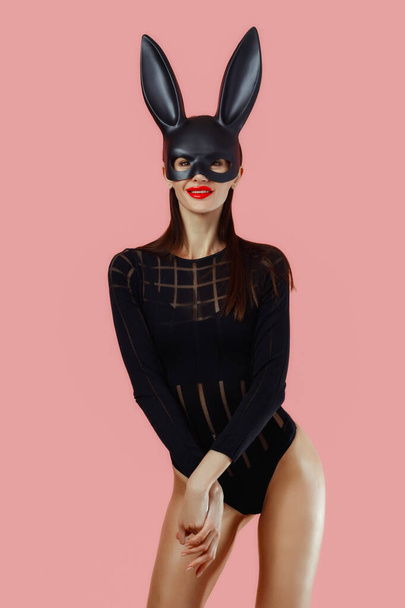 Sexy woman wearing a black mask Easter bunny standing on a pink background and looks very sensually. - Photo, Image