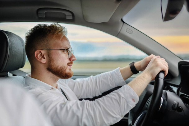 young male driver of a car in glasses and business clothes, rides at sunset. Business class driver concept - Фото, зображення