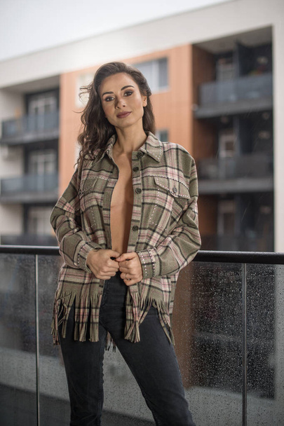 Gorgeous glamour brunette woman with plaid jacket posing on modern balcony with amazing view on city .Portrait of a Stylish fashionable woman with long legs , black jeans wearing in balcony - Valokuva, kuva