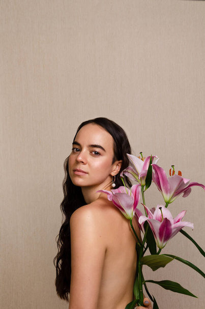 Portrait of a confident nude woman looking at camera, holding lilies flowers next to her back. Femininity, body image. - Фото, изображение