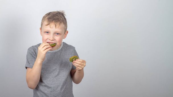 Un chico caucásico come un kiwi verde y ríe y sonríe sobre un fondo gris. - Foto, Imagen