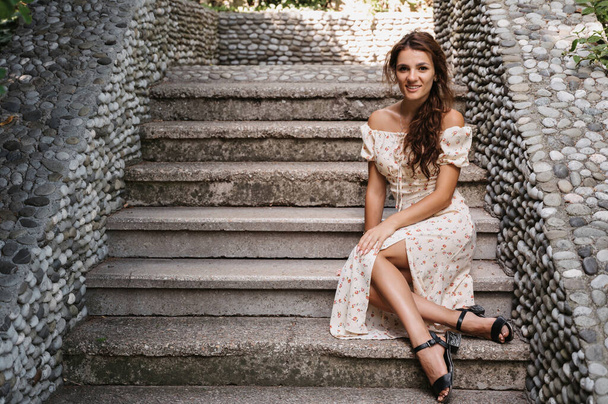 Una jovencita, morena de pelo largo, con un vestido vintage está sentada en los escalones de una escalera de piedra gris - Foto, Imagen