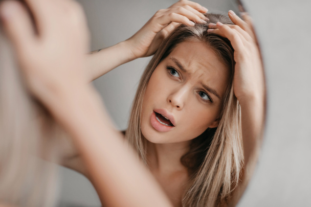 Femme frustrée cherchant des flocons de cheveux souffrant de problèmes de pellicules, regardant son reflet dans le miroir dans la salle de bain - Photo, image