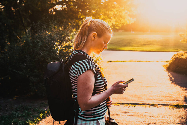 Gün batımında parkta akıllı telefon kullanan genç bir kadın. - Fotoğraf, Görsel