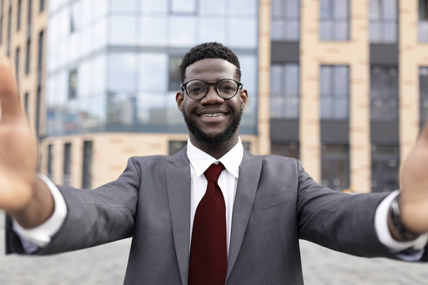 Feliz hombre de negocios afroamericano en traje formal tomando selfie mientras está parado fuera del centro de negocios, capturando momento - Foto, imagen