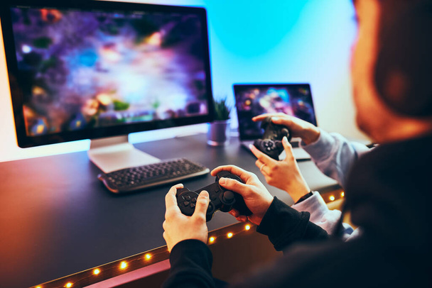 Friends playing video game at home. Gamers holding gamepads sitting at front of screen. Streamers girl and boy playing online in dark room lit by neon lights. Competition and having fun - Photo, Image