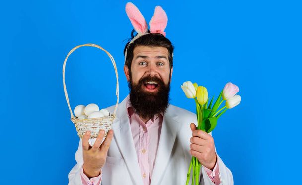 イースターの日。チューリップとバスケットの卵を持つウサギの耳のウサギの男。スーツを着た髭の男。春休み. - 写真・画像