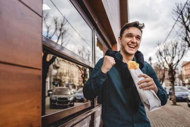 One young happy caucasian man standing outdoor with sandwich eating on the street happy celebrating success smiling real people copy space in spring or autumn or winter day - Φωτογραφία, εικόνα