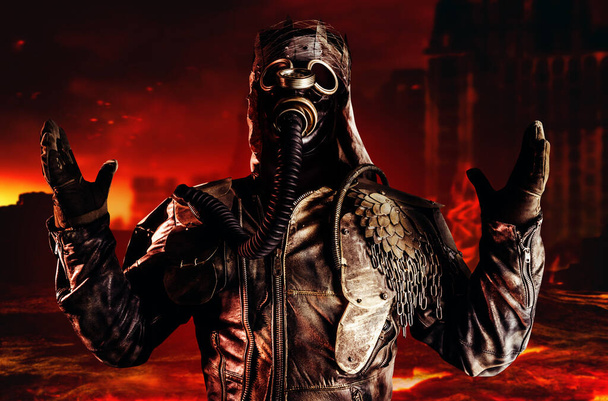 Photo of fantasy post apocalyptic warrior with armored outfit jacket, gas mask and scrap crown standing and praying on destroyed and burning city background. - Photo, Image