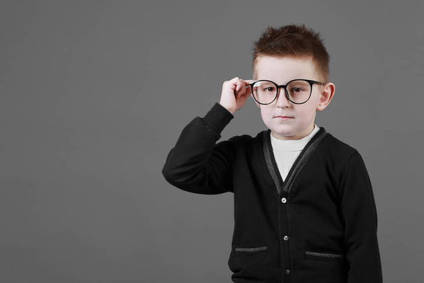 スマートチャイルドボーイは、グレーの背景、クローズアップに指で眼鏡を調整します。カジュアルな服のスタジオ少年の肖像画。コピースペース. - 写真・画像