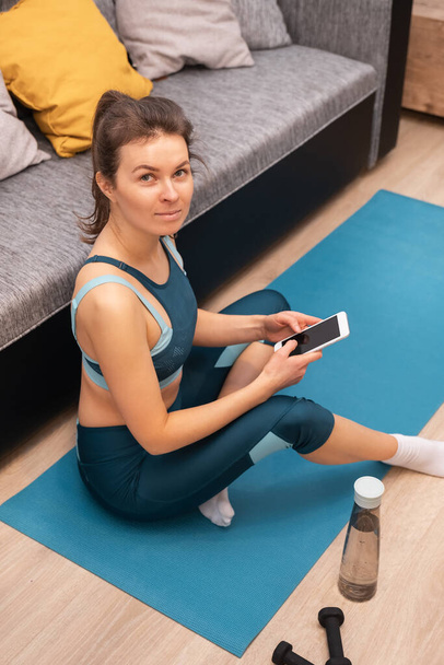 Giovane donna in abbigliamento sportivo guardando la fotocamera mentre seduto su un tappeto e tenendo in mano uno smartphone - Foto, immagini