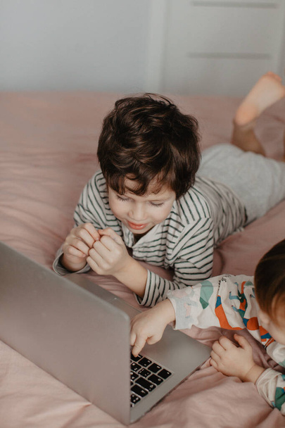 Little brother and sister lie in the bedroom on the bed and look at the laptop - Foto, imagen