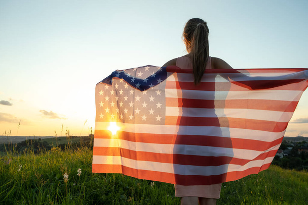 Vue arrière de la jeune femme heureuse posant avec le drapeau national des États-Unis debout à l'extérieur au coucher du soleil. Fille positive célébrant la fête de l'indépendance des États-Unis. Journée internationale de la démocratie concept - Photo, image