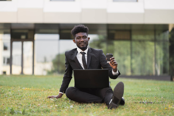 African businessman sitting on green grass outside office, showing thumb up while using laptop - Foto, Bild