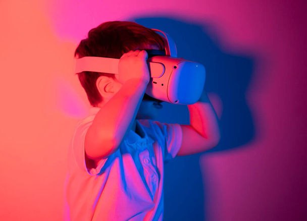 Junges Kind mit Virtual-Reality-Brille, kaukasischer Junge mit VR-Headset, spielt Videospiele auf Neon-Hintergrund  - Foto, Bild