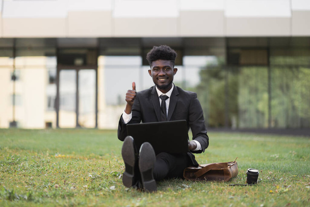 African-American man using laptop outdoors, sitting on the grass near office - Zdjęcie, obraz