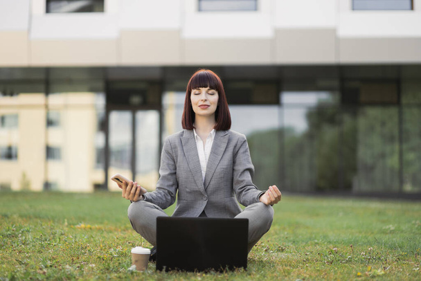 Pretty female executive practicing yoga, sitting with eyes closed outside office on green grass - 写真・画像