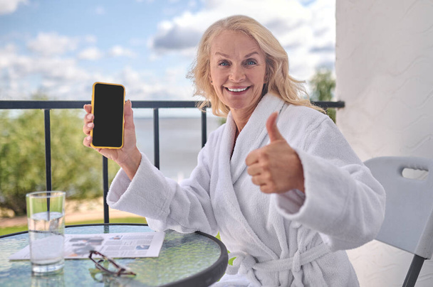 A blonde woman in a white robe with a smartphone in hands looking happy - Foto, Bild