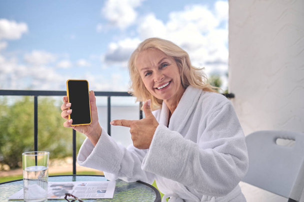 A blonde woman in a white robe with a smartphone in hands looking happy - Photo, image