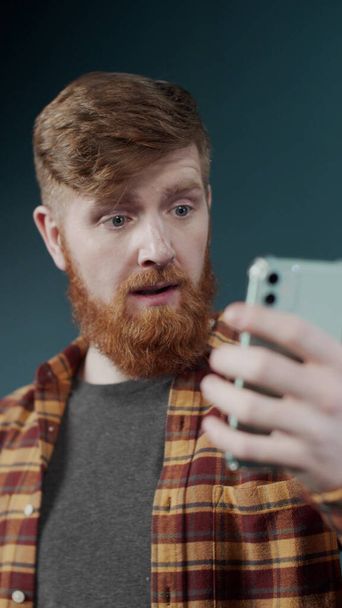Entusiástico jovem ruiva homem fazer uma chamada de vídeo por câmera de telefone perto de uma parede verde - Foto, Imagem