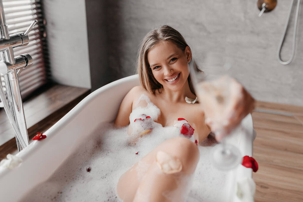 gelukkig vrouw drinken champagne terwijl liggend in schuimige badkuip, vieren vakantie en stretching glas naar camera - Foto, afbeelding