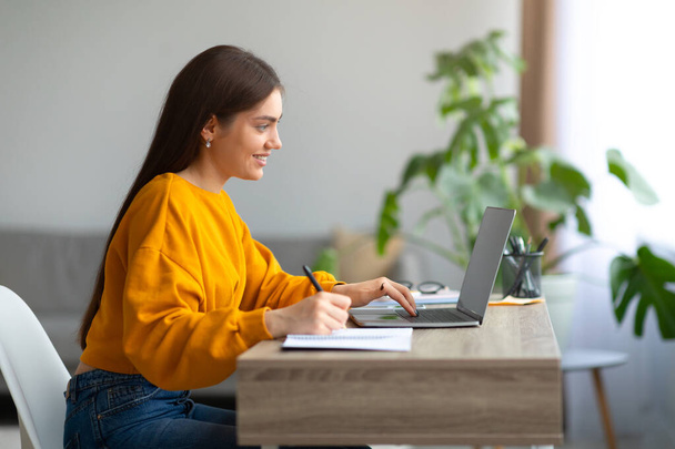 Positieve jonge vrouw schrijven in notebook tijdens online lezing op laptop, communiceren met collega 's op webcam - Foto, afbeelding