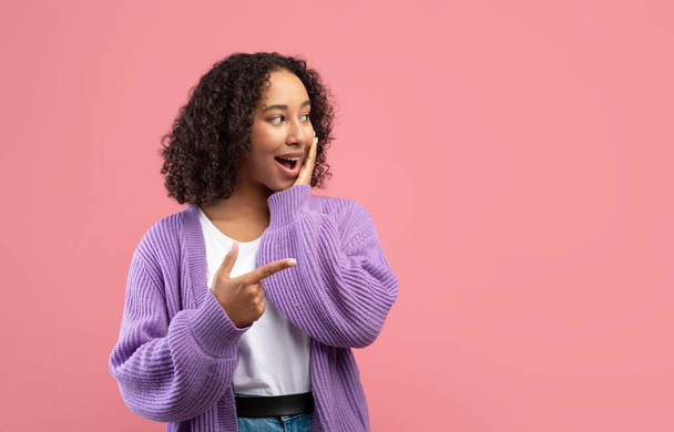 Opgewonden jonge zwarte dame wijzen naar lege ruimte, verbaasd over enorme verkoop over roze studio achtergrond - Foto, afbeelding