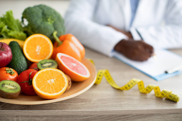 Здравоохранение и диета. Женский диетолог пишет план диеты, сидит за столом с овощами и фруктами на тарелке - Фото, изображение