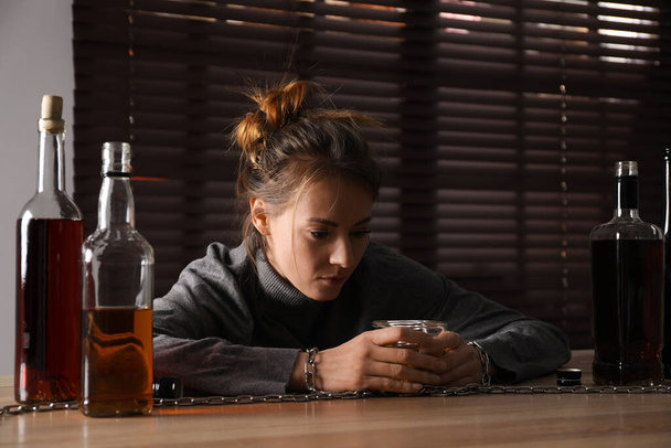 Alkoholfüggőség. Nő láncra verve egy üveg likőrrel a fa asztalnál a szobában. - Fotó, kép