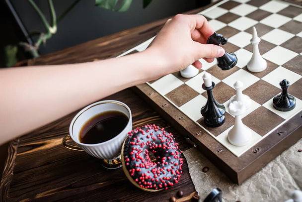 Die Hand hält eine weiße Schachfigur. Schachbrett und Streuschach auf Holztisch. Schokokrapfen zum Frühstück am Morgen. Brettspielkonzept - Foto, Bild