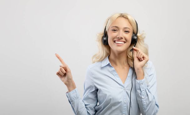 Ευτυχισμένη νεαρή γυναίκα τηλεφωνήτρια φορώντας ακουστικά και δείχνοντας στην άκρη τον ελεύθερο χώρο, το φως φόντο, πανόραμα - Φωτογραφία, εικόνα