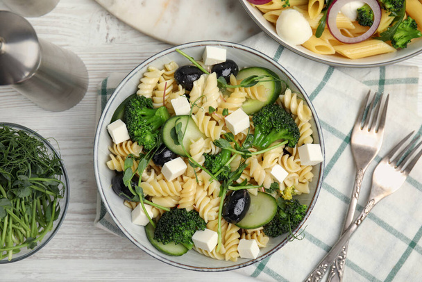Bowl of delicious pasta with cucumber, olives, broccoli and cheese on white wooden table, flat lay - Valokuva, kuva