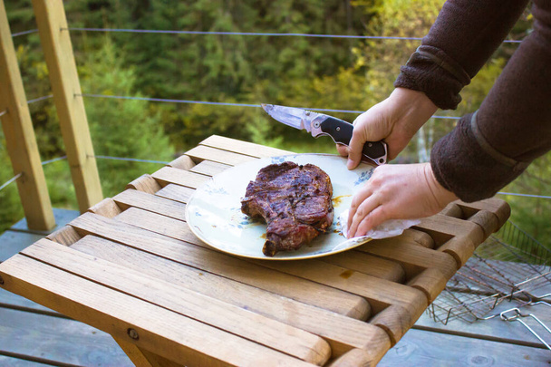 Freshly roasted bbq meat lies on plate standing on a wooden table. Female hand holding a knife, preparing to cut a steak. A picnic outdoors in a green pine forest. Eco-tourism, relaxing on the nature. - Photo, Image