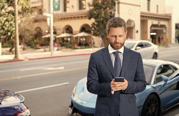 guapo hombre de negocios charlando en el teléfono inteligente stand by auto de lujo al aire libre. coche disponible. - Foto, imagen