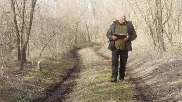  un hombre camina a lo largo de un camino forestal con hierba y mira en su computadora portátil transformador - Metraje, vídeo