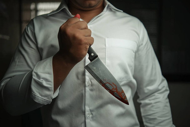 Um homem de negócios criminoso usando uma camisa branca de mangas compridas segurando uma faca sangrenta está agredindo brutalmente sua vítima em uma sala, há espaço para texto, Conceito Criminal, Crime, Assassinato. - Foto, Imagem