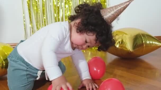 Boldog vegyes fajú kisgyerek ül a földön a háttérben egy díszített szobában a születésnapján. - Felvétel, videó