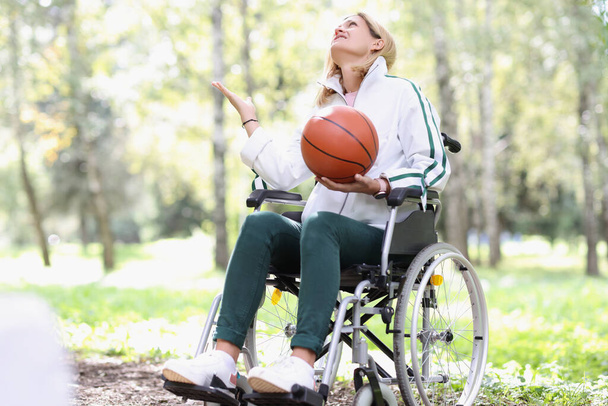 Νεαρή γυναίκα κάθεται σε αναπηρική καρέκλα κρατώντας μπάσκετ στο πάρκο - Φωτογραφία, εικόνα