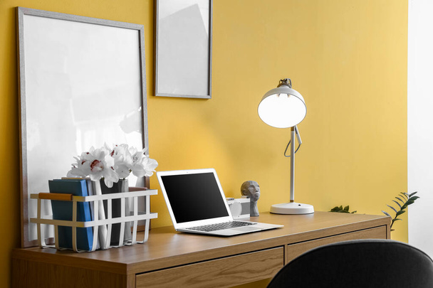 ラップトップ、フレーム、黄色の壁の近くに光るランプのあるモダンな職場 - 写真・画像
