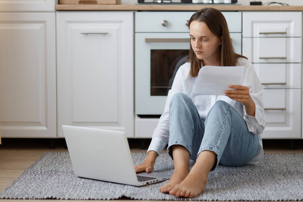 Indoor shot of concentrated young adult woman wearing white shirt and jeans sitting on floor in kitchen and working on notebook, paying utility bills, looking at laptop screen. - Fotografie, Obrázek