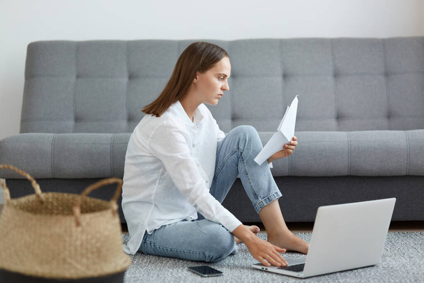 Horizontal shot of dark haired serious woman wearing white shirt and jeans sitting on floor with gray sofa on background, reading information on paper, working on laptop. - Photo, Image