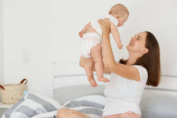 indoor shot of satisfied smiling woman wearing white shirt and short sitting on bed with her daughter, mother throwing up her toddler kid, playing with child, looking with love and gentle. - Foto, immagini