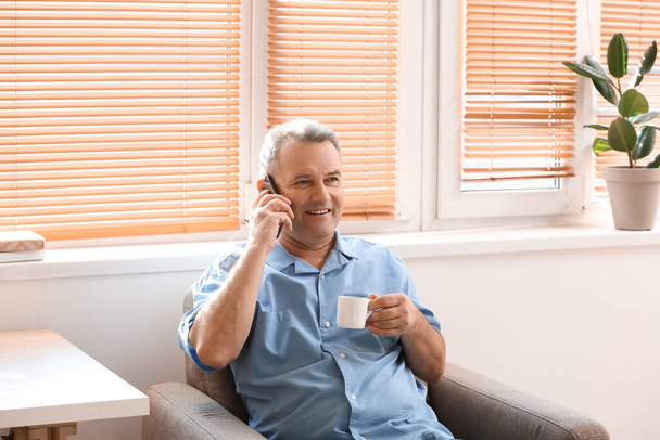 Senior man with cup of coffee talking by mobile phone in armchair at home - Foto, Imagem