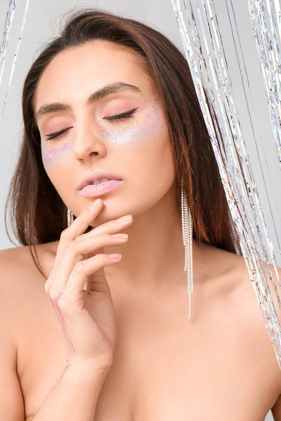 Beautiful young woman with glitters on her face against light background - Photo, Image