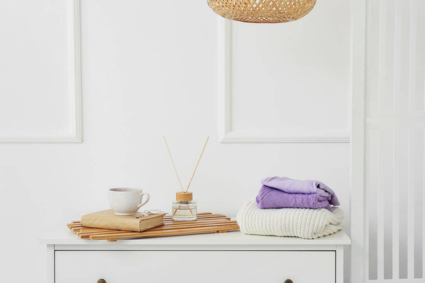 Складные свитера, тростниковый диффузер, чашка и книга на комоде возле светлой стены - Фото, изображение