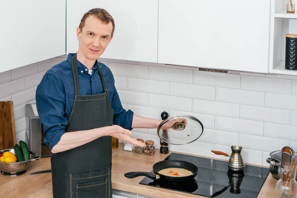 Mężczyzna w średnim wieku z krótkimi rudymi włosami w czarnym fartuchu stojący trzymając szklaną pokrywę patelni, pokazując świeży omlet. - Zdjęcie, obraz