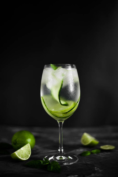 A wine glass of fresh cold lemonade with ice, cucumber slices and lime fruit on  the dark background - Фото, зображення