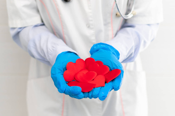 Τα χέρια ενός γιατρού με λευκό ιατρικό παλτό και λαστιχένια γάντια με φωνοσκόπιο κρατούν πολλές κόκκινες καρδιές στο νοσοκομείο. Επιλεκτική εστίαση. Κοντινό πλάνο - Φωτογραφία, εικόνα