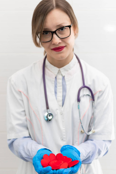 Η νεαρή όμορφη γιατρός με το λευκό ιατρικό παλτό και τα λαστιχένια γάντια με το φωνοενδοσκόπιο κρατά μια κόκκινη καρδιά στα χέρια της στο νοσοκομείο. Επιλεκτική εστίαση. Πορτρέτο - Φωτογραφία, εικόνα