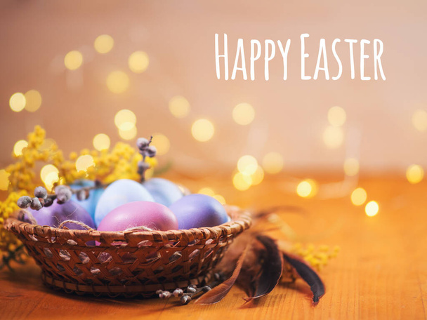 Happy Easter holiday card, pastel colored eggs in a wicker basket close-up, willow and mimosa branch - Foto, Imagen